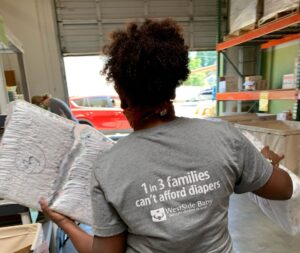 WestSide Baby staff member carrying diapers in the warehouse, wearing a grey t-shirt that reads: "1 in 3 families cant afford diapers"